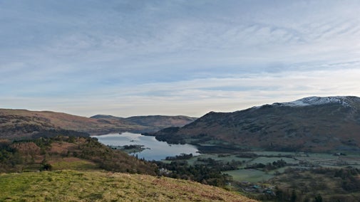 View of Ullswater and surrounding hills just after sunrise on an early spring day above the Patterdale Circular walking path. The sky is light blue with wispy clouds, the lake is still, and the fell tops have a dusting of snow. The valley floor is still in the shadow of the hills above.